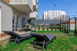 A modern outdoor seating area with lounge chairs and a table, surrounded by a well-maintained lawn and a building with multiple windows. - Historic City Stay for 4 in Warsaw (Łódź)