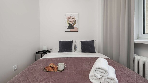 Uma cama bem arrumada com cobertura acolchoada, uma bandeja com croissants e uma chávena, e uma toalha enrolada ao lado.