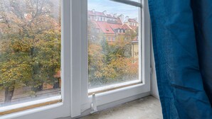 Ikkunasta avautuu näkymä rakennuksiin ja puihin, verho on osittain vedettynä ja ikkunan karmi on valkoinen.