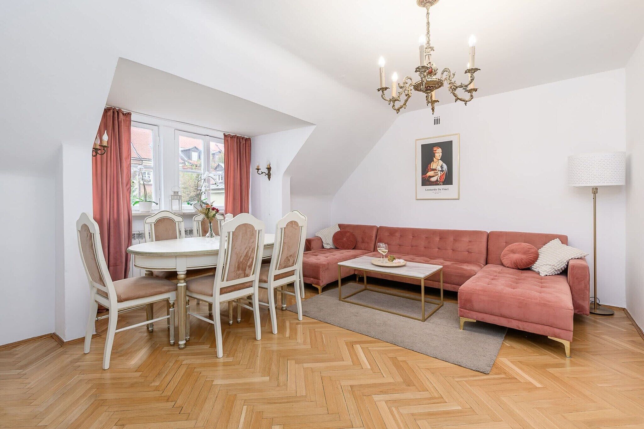 Un coin repas avec une table ronde et des chaises, un canapé rose, un lustre et une œuvre d’art encadrée accrochée au mur.