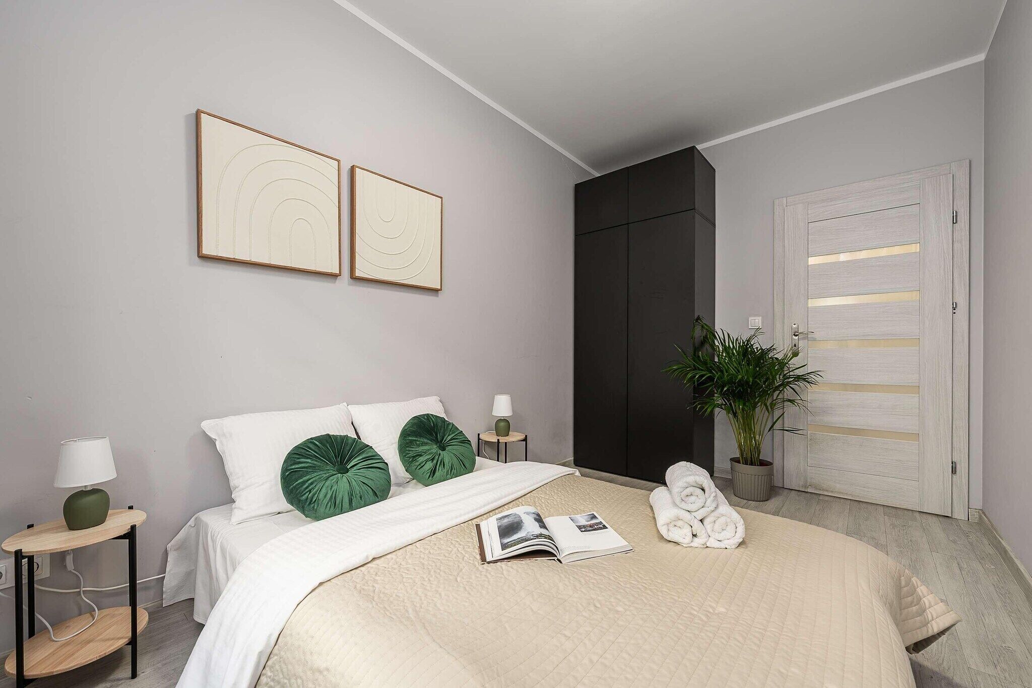 Un dormitorio con cama, mesita de noche, lámpara, planta en maceta y puerta con luminaria.