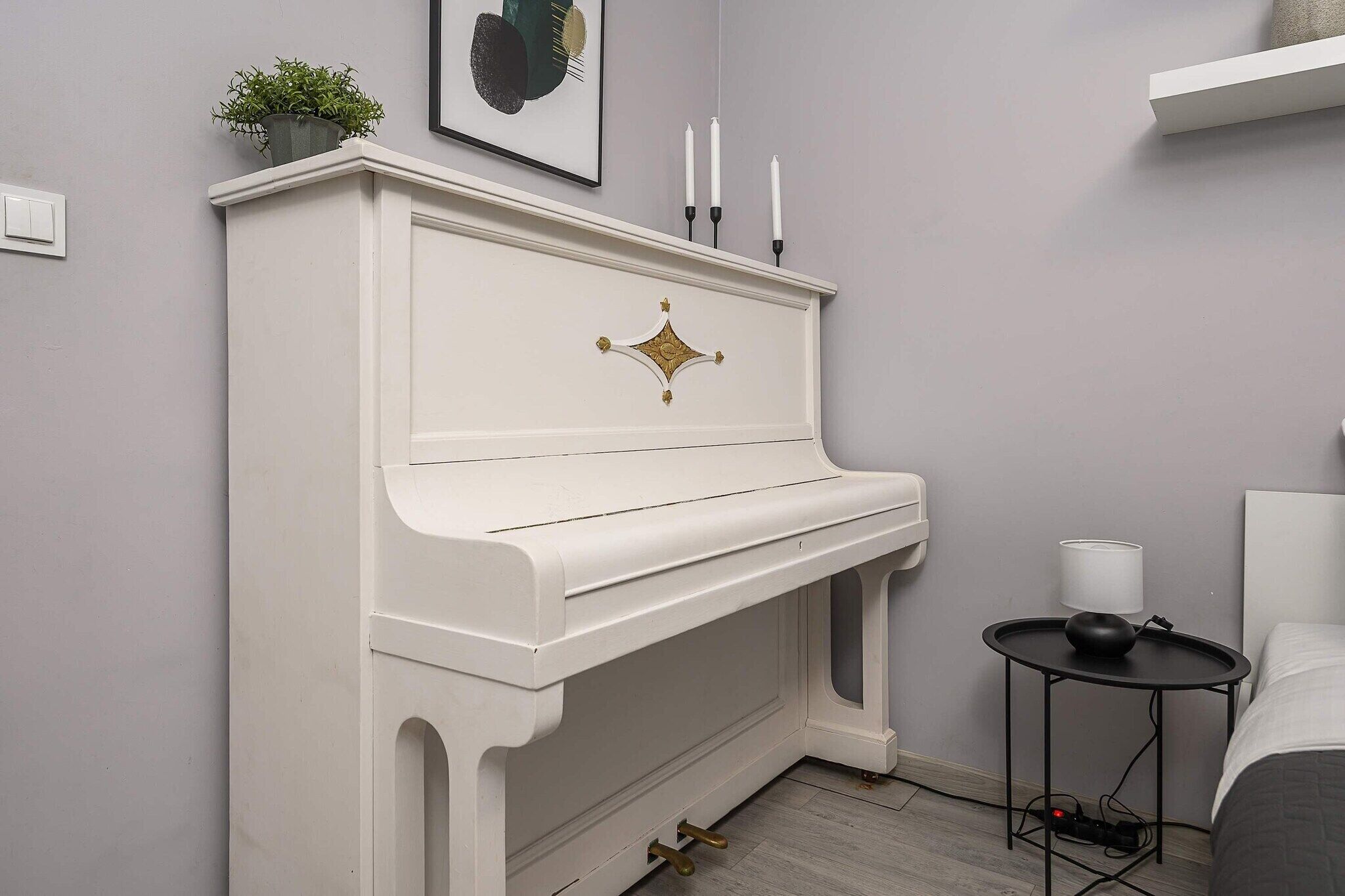 Ein weißes Klavier mit goldfarbenen Akzenten, ein kleiner runder Tisch mit schwarzem Fuß und eine weiße Lampe an einer grauen Wand.