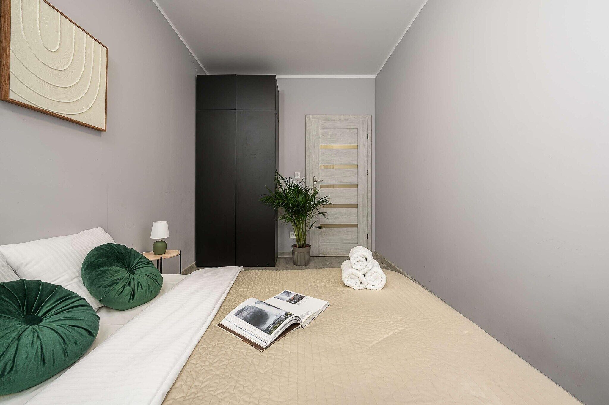 Un dormitorio con cama, almohadas, una mesita de noche con lámpara, una planta y una puerta con perchero para toallas.