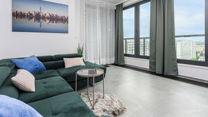 Ett modernt vardagsrum med en grön soffa, en bärbar dator på ett litet bord och utsikt över stadsbilden genom stora fönster.
