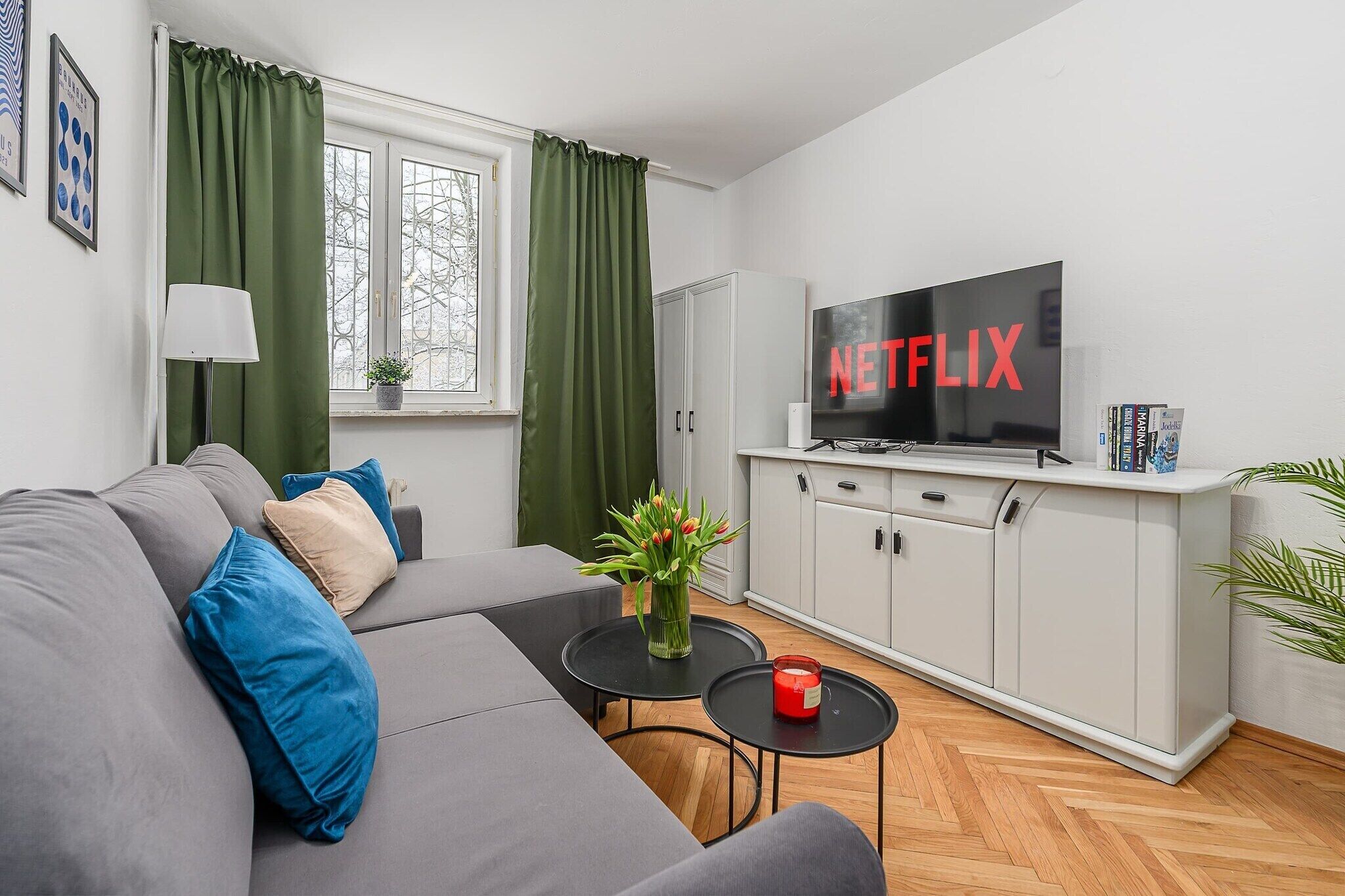 En moderne stue med en grå sofa, et fladskærms-tv, et skab og et lille bord med en vase og et lys.