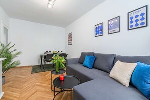 Ein modernes Wohnzimmer mit einem grauen Sofa, einem kleinen runden Tisch mit einer roten Kerze und einem Essbereich mit einem für zwei Personen gedecktem Tisch.