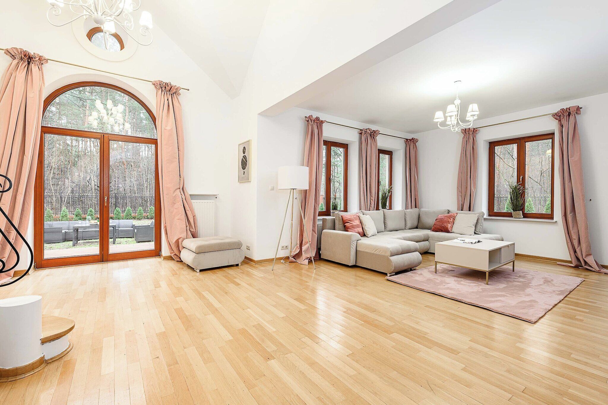 A spacious living room with a large sofa set, a coffee table, and a floor lamp. There are large windows with curtains, a view of the outdoors, and a wooden floor.