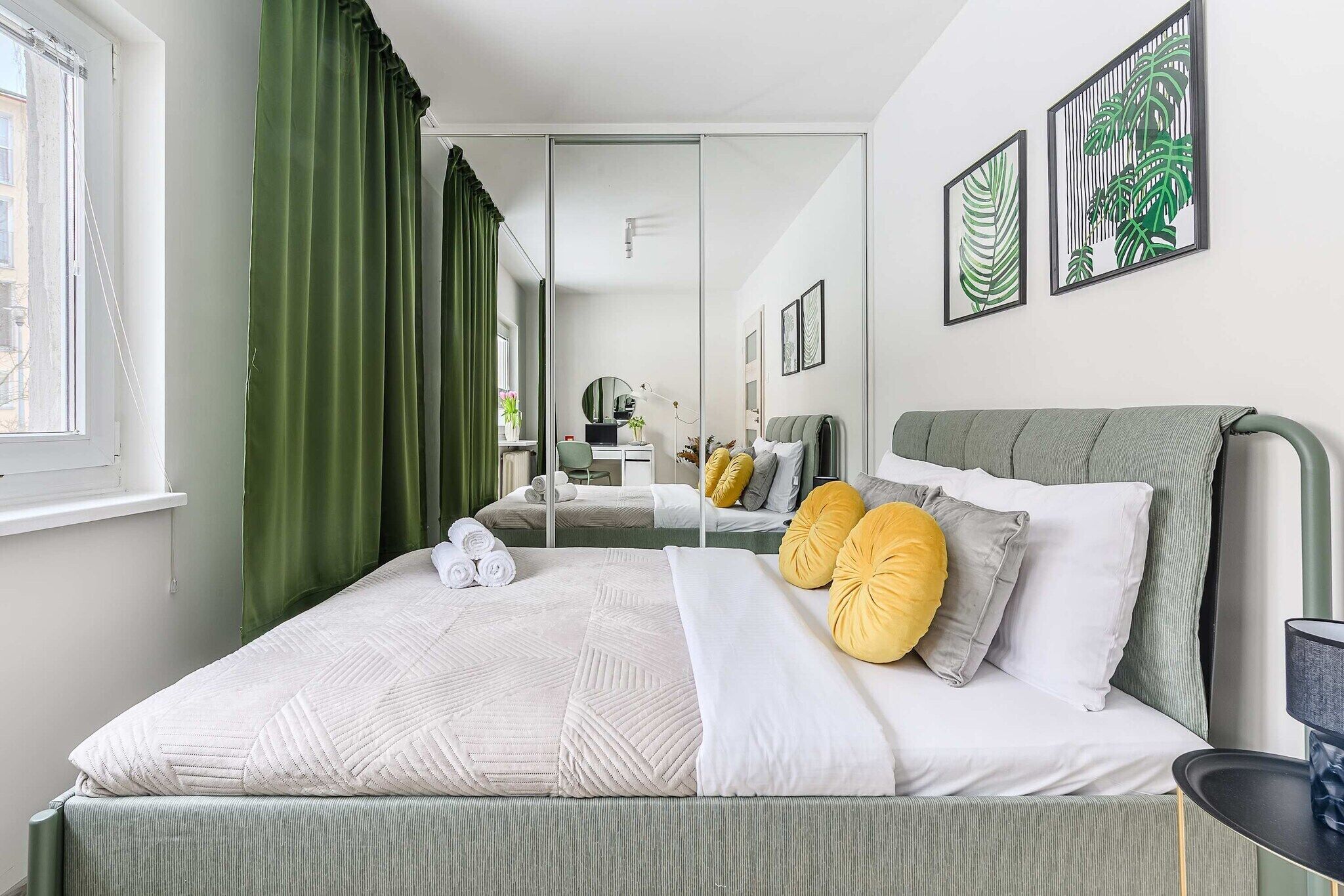 Une chambre moderne avec un lit, des rideaux verts, un miroir et des œuvres d’art accrochées au mur.