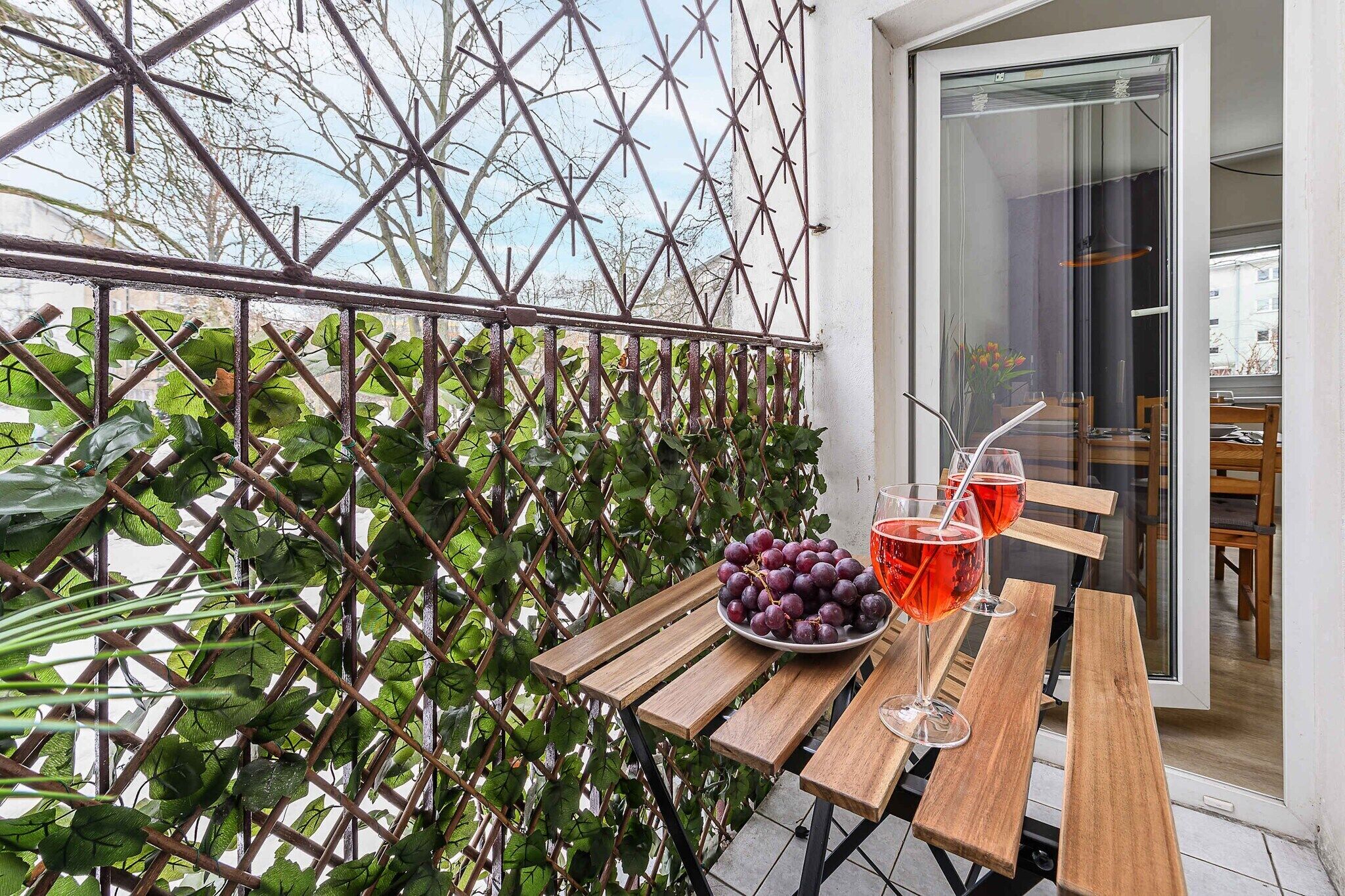 A wooden table with a bowl of grapes and two glasses of red beverage, a metal trellis with green foliage, and a glass door leading to an interior space.