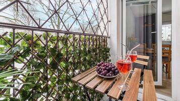 A wooden table with a bowl of grapes and two glasses of red beverage, a metal trellis with green foliage, and a glass door leading to an interior space.