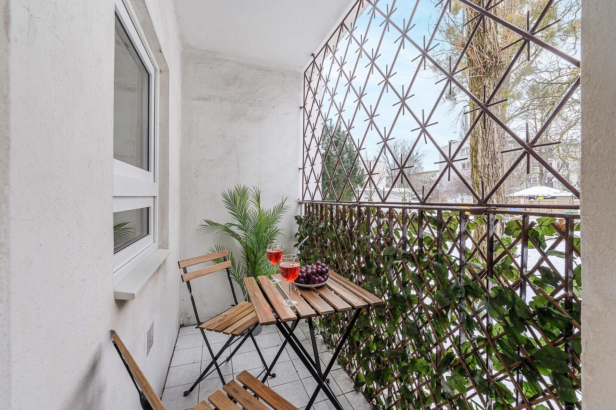 Een klein balkon met een houten tafel, twee stoelen, een bloempot en een metalen hek.