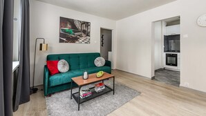 A living room with a teal sofa, a coffee table with books, a painting on the wall, and a kitchen in the background. - Warsay Apartment near Metro (Warszawa 2)