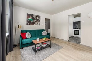 A living room with a teal sofa, a coffee table with books, a painting on the wall, and a kitchen in the background. - Warsay Apartment near Metro (Warszawa 2)