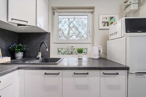 A modern kitchen with a black countertop, white cabinets, a stainless steel sink, a window with a geometric pattern, a white kettle, and a microwave. - Warsay Apartment near Metro (Warszawa 2)