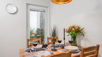 A dining area with a table set for four, a clock on the wall, and a window with a view of greenery.