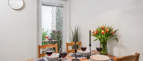 A dining area with a table set for four, a clock on the wall, and a window with a view of greenery.