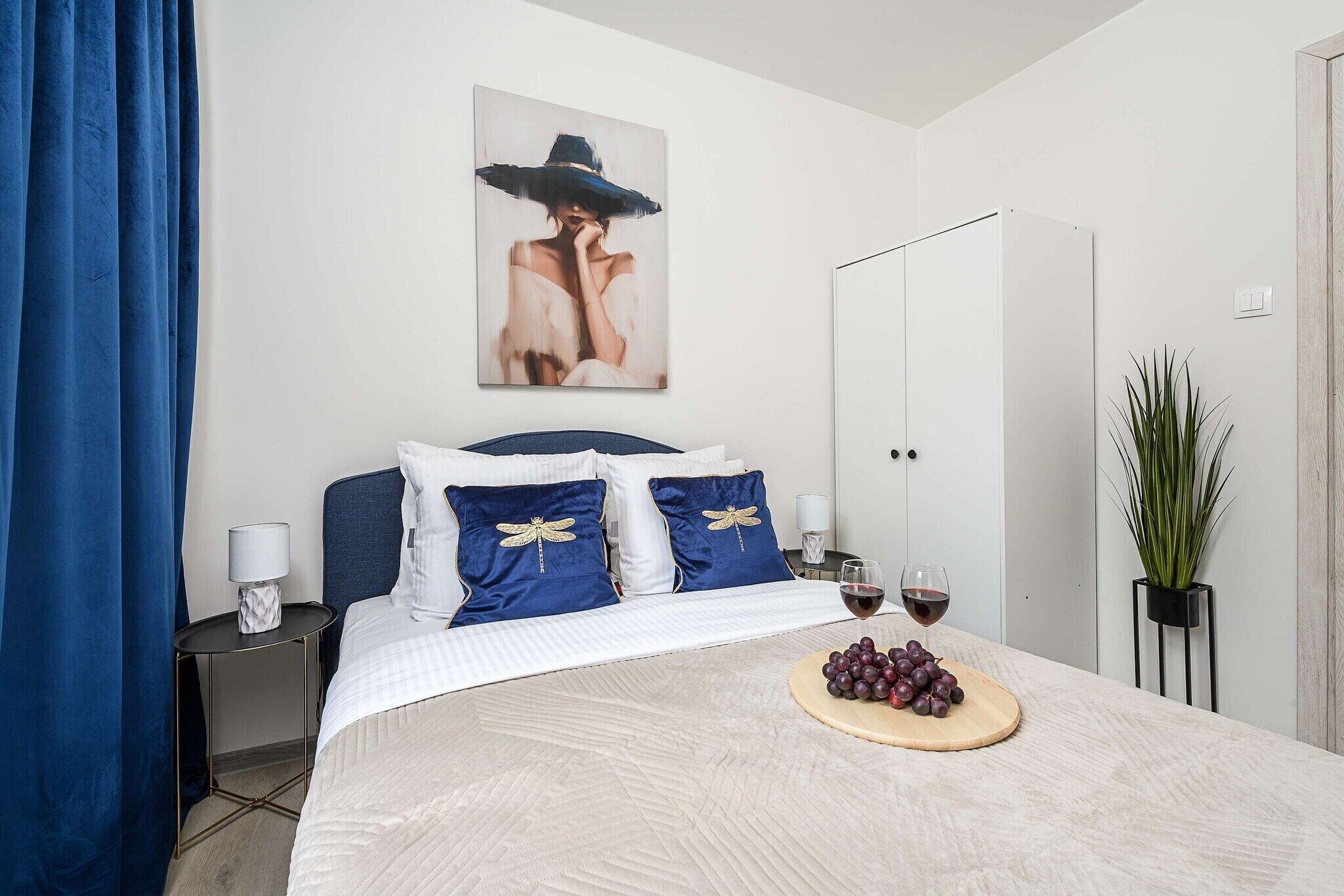 A neatly arranged bedroom with a bed, pillows, a bedside table, a nightstand, a wardrobe, a potted plant, and a fruit platter with wine glasses.