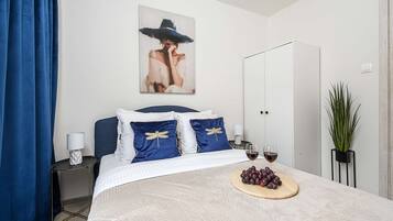 A neatly arranged bedroom with a bed, pillows, a bedside table, a nightstand, a wardrobe, a potted plant, and a fruit platter with wine glasses.