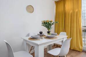 A dining area with a white table set for two, a clock on the wall, and a yellow curtain. - Warsaw Flat on Pereca (Warszawa 2)