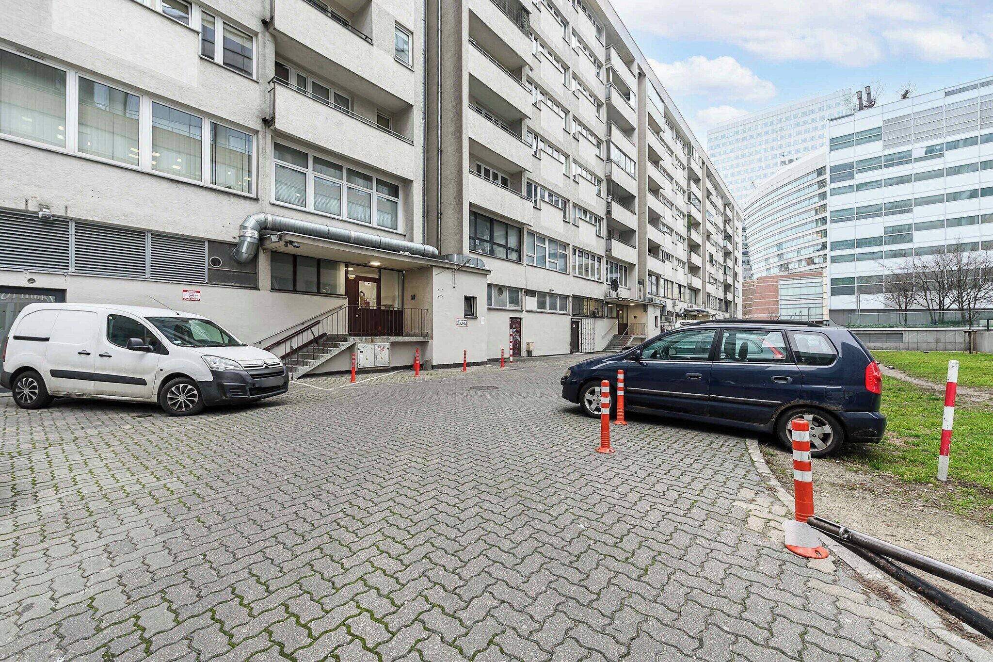 Un immeuble à plusieurs étages avec un espace de stationnement, un fourgon blanc et une voiture bleue garés dans la rue.