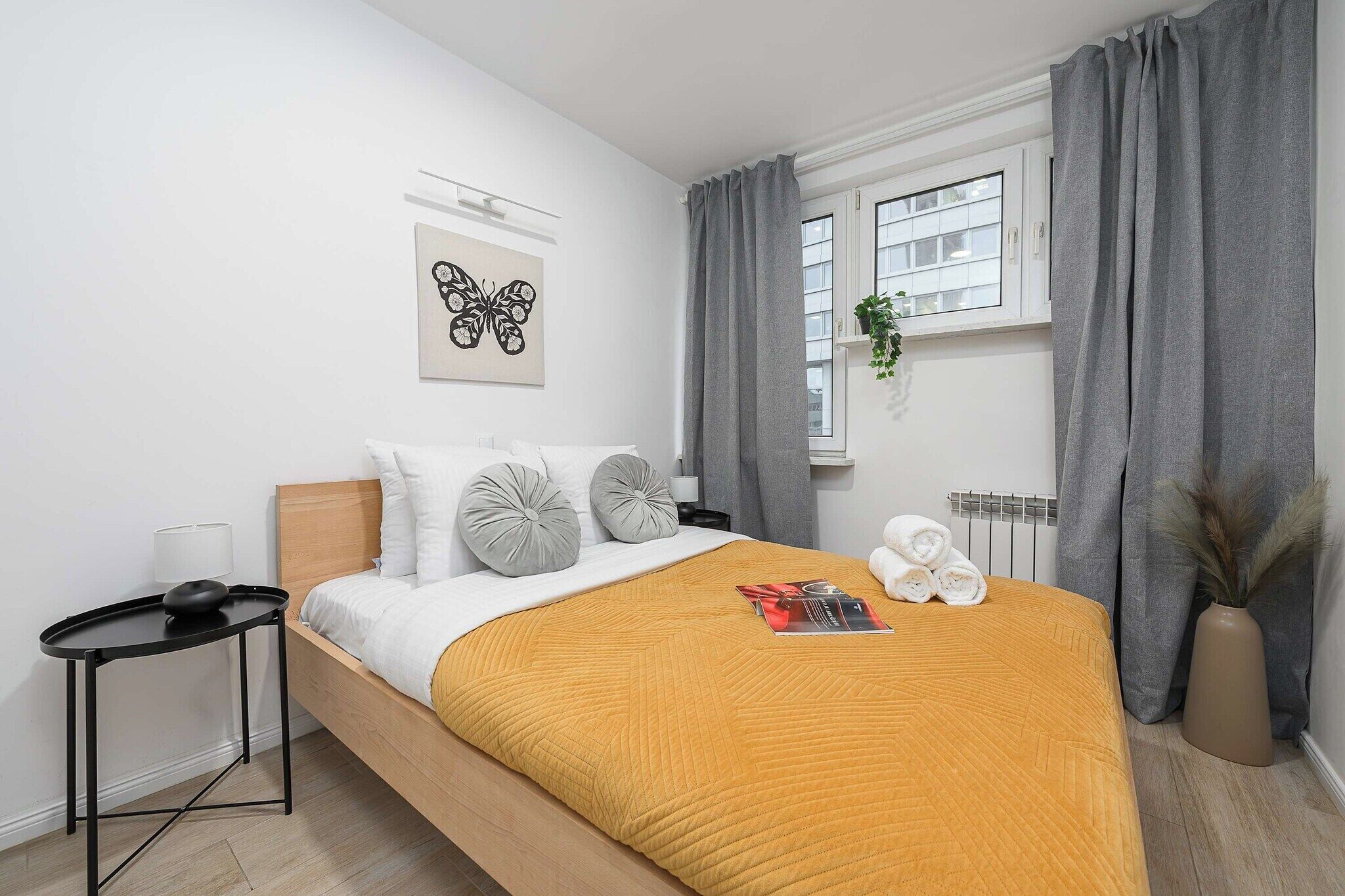 Une chambre moderne avec un lit en bois, une table de chevet, une lampe fixée au mur, une fenêtre avec des rideaux et une œuvre d’art représentant un papillon, encadrée.