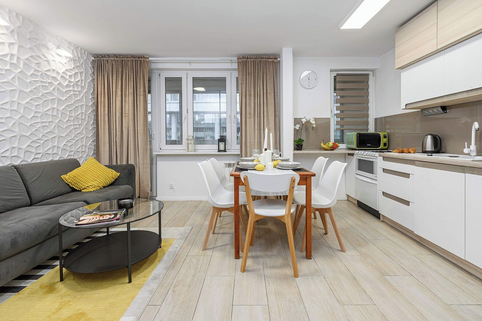 Uma sala de estar moderna com um sofá cinzento, uma mesa de centro de vidro e um espaço de jantar com cadeiras brancas e uma mesa de madeira.