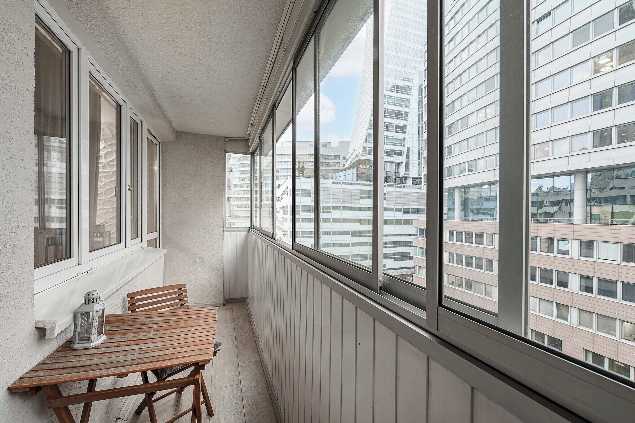 A balcony with a wooden table and chairs, large windows, and a view of modern buildings.