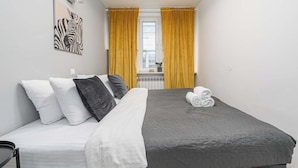 Ein Einzelbett mit grauer Bettdecke, weißen und schwarzen Kissen sowie einem zusammengerollten Handtuch seitlich.