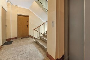 A hallway with a wooden door, a staircase with a metal railing, and a wall-mounted light switch. - Warsaw Flat with Church Views (Warszawa 2)