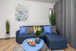 Ein modernes Wohnzimmer mit einer Eckcouch, einem Couchtisch mit Gebäck, einer Vase mit Blumen und einem großen abstrakten Gemälde an der Wand.