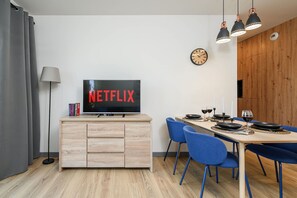 Ein modernes Wohnzimmer mit Fernseher, der 'NETFLIX' anzeigt, eine Essecke mit blauen Stühlen und eine Holzkommode.