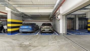 A parking garage with two cars parked in designated spaces, concrete pillars with yellow and black stripes, and a circular traffic sign on the wall.