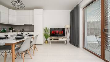 Un salon moderne avec un téléviseur à écran plat affichant le logo de Netflix, un coin repas avec une table dressée pour deux personnes et une grande fenêtre équipée de stores.