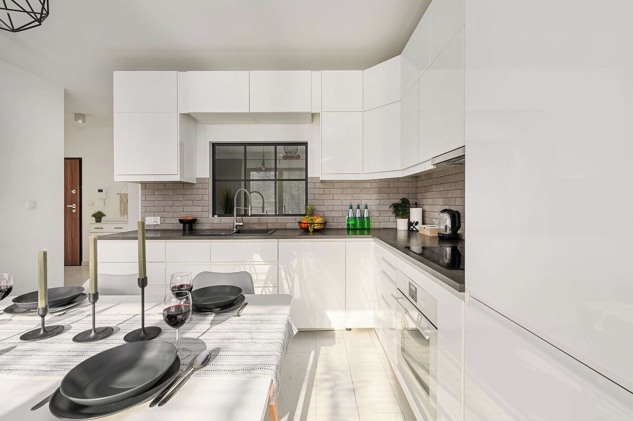 A modern kitchen with white cabinetry, a black countertop, and a dining area set for two with wine glasses and plates.