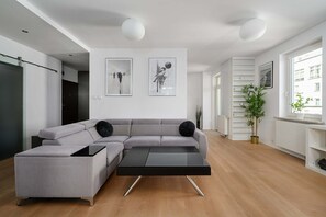 Ein modernes Wohnzimmer mit einem grauen Ecksofa, einem schwarzen Couchtisch und Holzboden. Zwei Wandleuchten und zwei gerahmte Kunstwerke sind über der Couch angebracht.