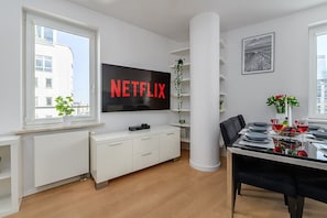 Ein modernes Wohnzimmer mit Fernseher, der 'NETFLIX' anzeigt, einem Essbereich mit Tisch für vier Personen und einem großen Fenster mit Blick auf Gebäude.