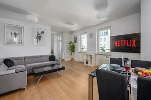 Een moderne woonkamer met een grijze bank, een zwarte salontafel en een eethoek met een schaal fruit en wijnglazen.