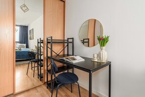 Ein Zimmer mit Holzboden, einem schwarzen Metallschreibtisch mit Stuhl, einem runden Spiegel und einer Vase mit Blumen.