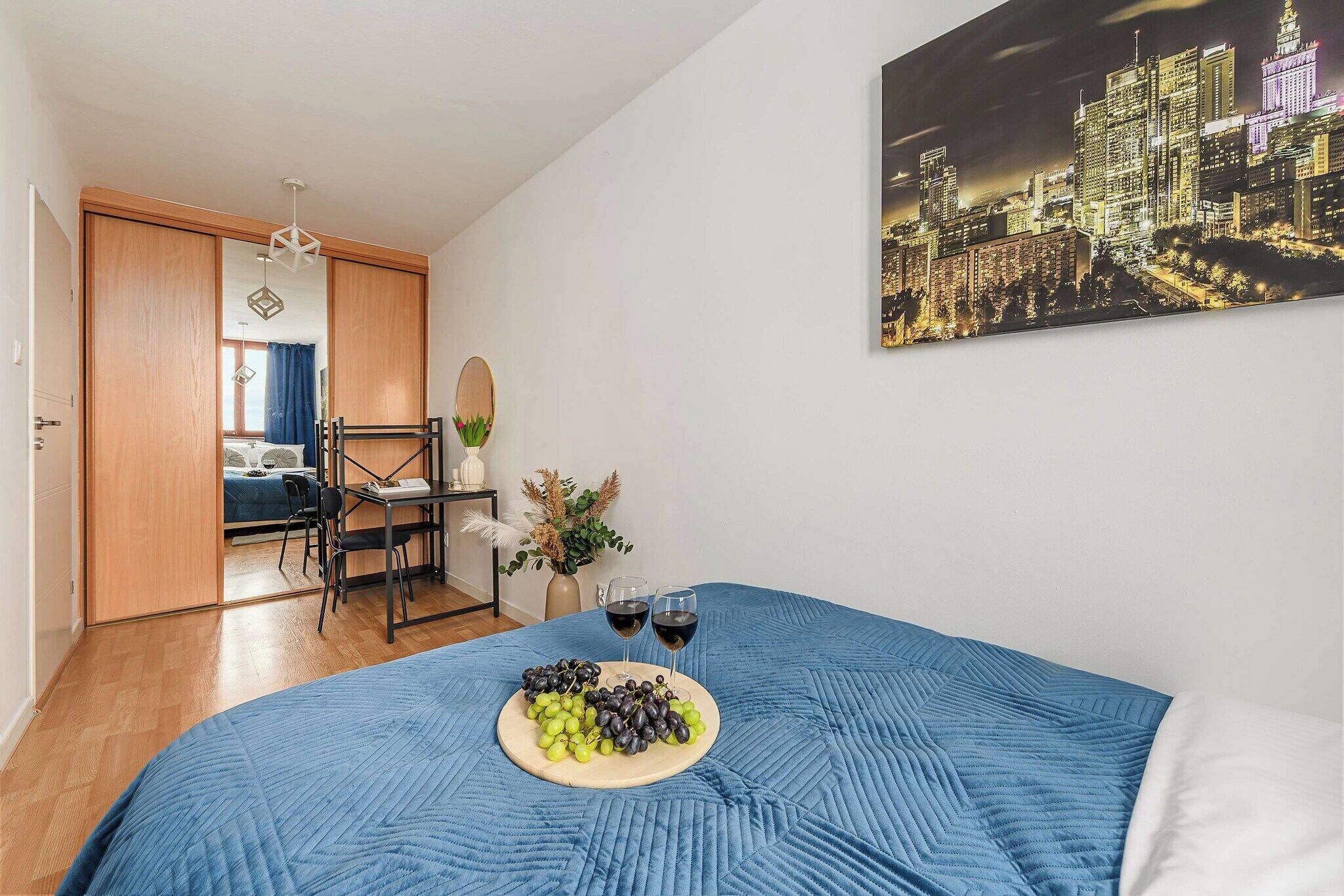 Une chambre à coucher comprenant un lit, un bureau, une chaise, une table de chevet, un vase avec des fleurs et un tableau représentant un paysage urbain accroché au mur.