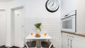 Ett modernt kök med vita skåp, vita kakelplattor ovanför arbetsbänken, en väggklocka, ett vitt bord med stolar, en vas med blommor och en frukostuppsättning med croissanter och apelsinjuice.