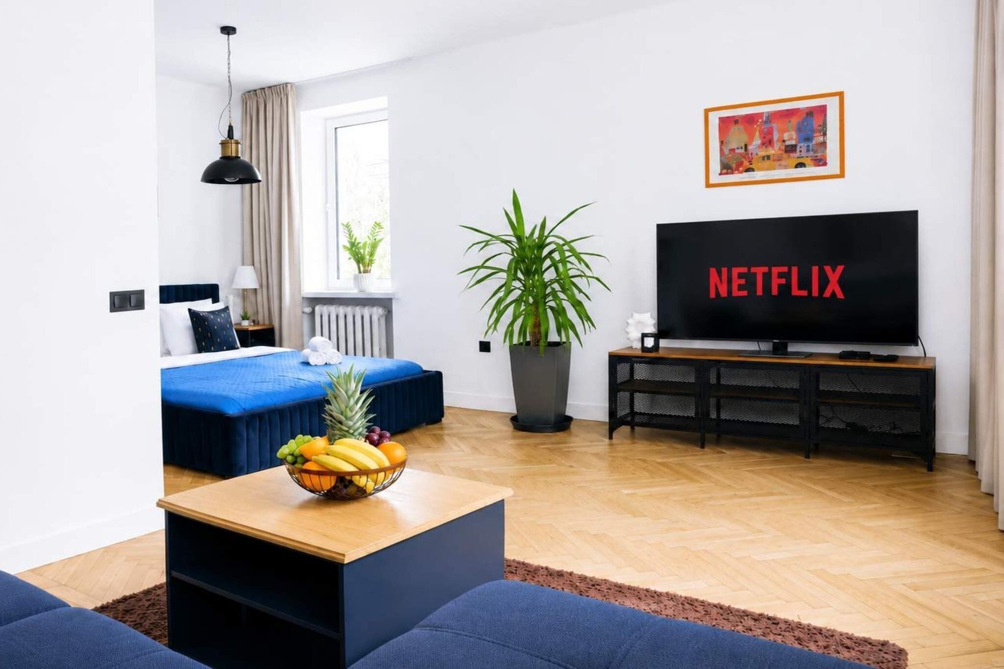 A modern living room with a bed, a TV, a fruit bowl, and a potted plant.