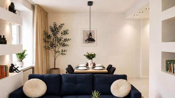 Ein modernes Wohnzimmer mit einem dunkelblauen Sofa, einem Couchtisch mit Obst, einem Essbereich mit gedecktem Tisch für zwei Personen und einer großen Pflanze.