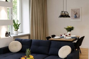 Ein modernes Wohnzimmer mit einem dunkelblauen Sofa, einem Esstisch für vier Personen und einem großen Fenster mit Vorhängen.