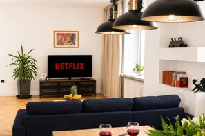 Ein modernes Wohnzimmer mit einem dunkelblauen Sofa, einem Flachbildfernseher, der 'NETFLIX' anzeigt, einem Couchtisch mit Gläsern und einer Topfpflanze.