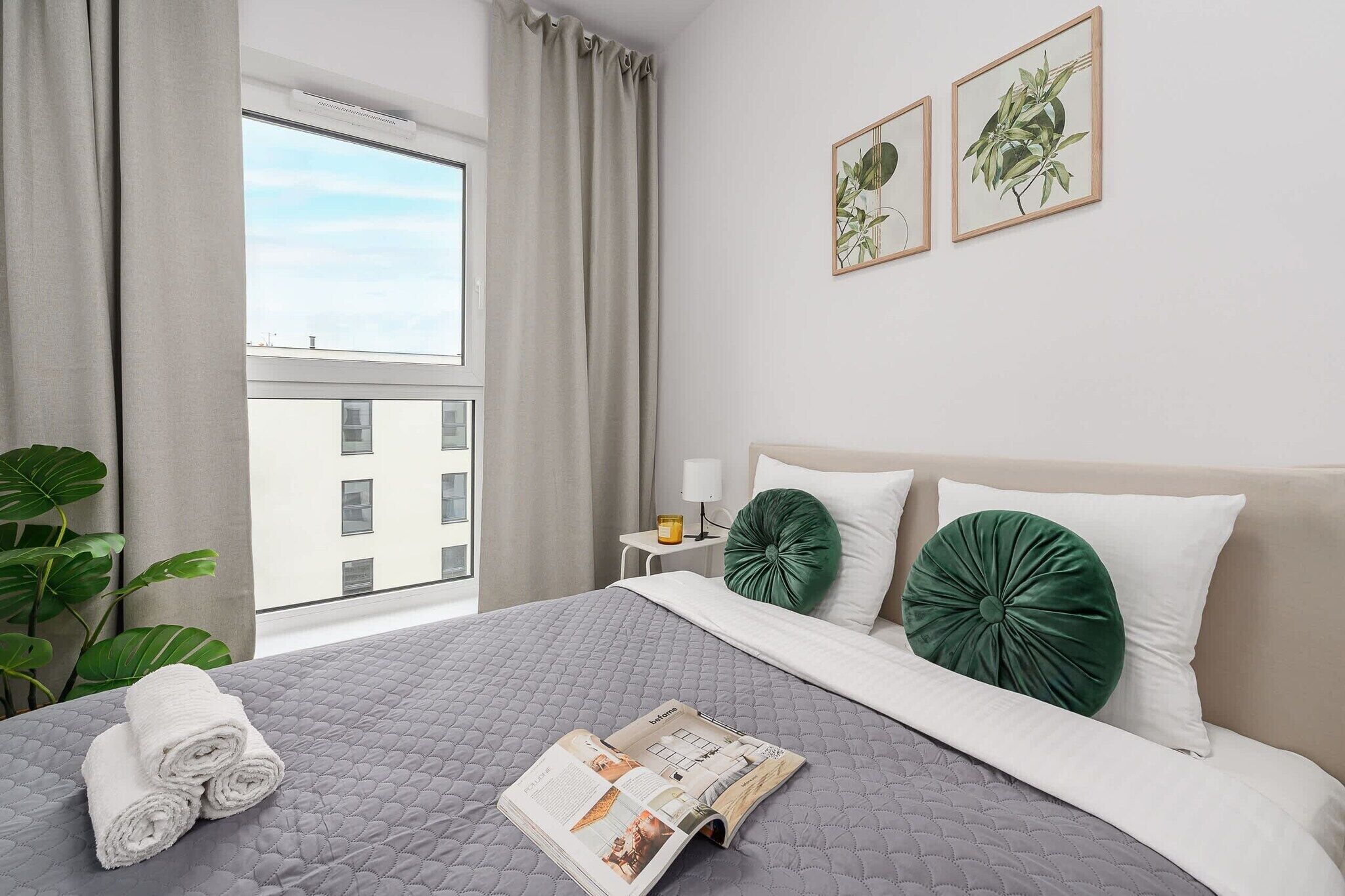 Une chambre d’hôtel avec un grand lit, une fenêtre avec des rideaux, une table de chevet avec une lampe et deux reproductions botaniques encadrées accrochées au mur.