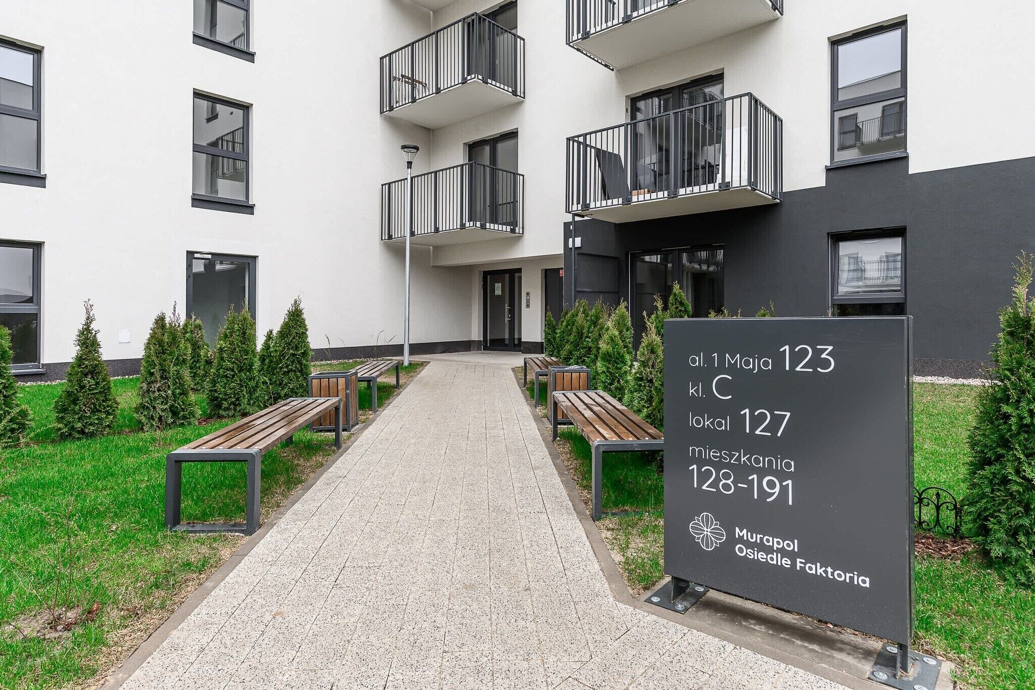 A modern building with a paved entrance, benches, and a sign indicating room numbers and location.