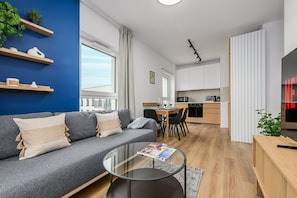 A modern apartment with a grey sofa, a glass coffee table, a kitchen with white cabinets, and a blue accent wall. - Łódź City Apartment with Balcony (Łódź)
