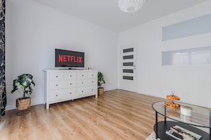 Ein modernes Wohnzimmer mit Fernseher, der 'NETFLIX' anzeigt, einem weißen Schrank mit Schubladen, zwei Topfpflanzen und einem Glastisch mit Gebäck.
