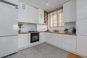 A modern kitchen with white cabinetry, a gas stove, an oven, and a window with blinds. - Family Apartment near Vistula River (Warszawa 2)