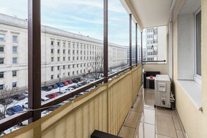 Ein Balkon mit Blick auf eine Stadtstraße und geparkte Autos, ein kleiner Tisch mit einem roten Gegenstand und ein weißes Gerät.